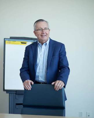 John Morris in a jacket standing with a chair